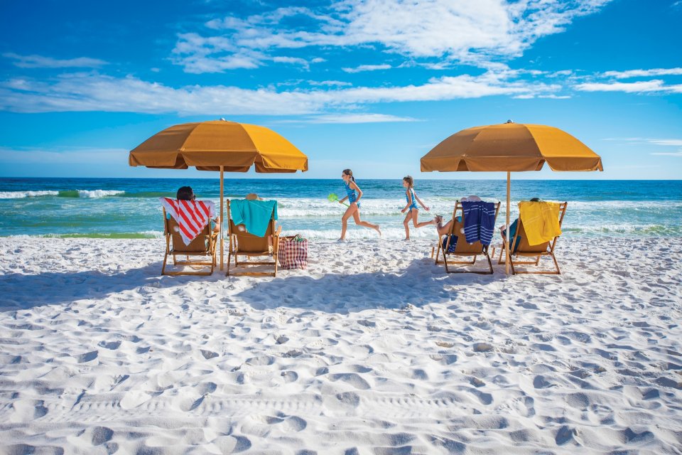Families sit on chairs beneath umbrellas on the white sand of a Pensacola beach as children run along the shore.