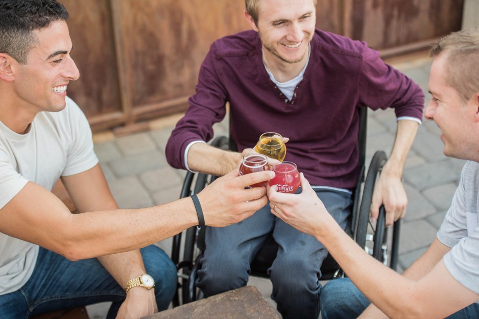 A group, including a man in a wheelchair, sits around an outdoor table and toasts with glasses of beer.