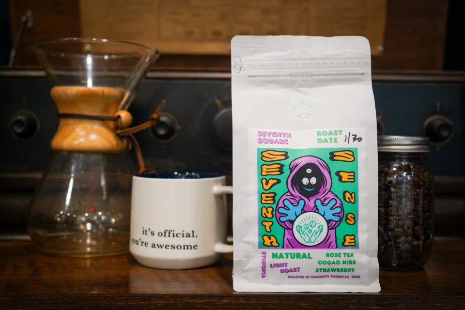 White coffee bag with colorful artwork and the words "Seventh Sense" on a table with a mug, Chemex carafe, and jar of beans.